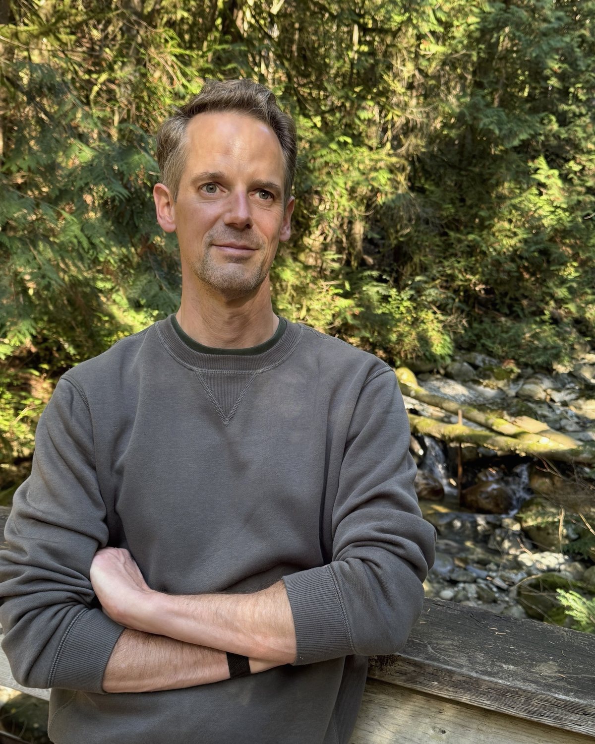 Sebastian Schilling in a grey sweater, arms crossed, leaning on a wooden railing above a forest creek in Lynn Canyon